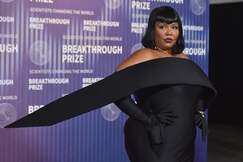 Lizzo poses with her hands on her hips in a black gown against a purple backdrop at the 2024 Breakthrough Prize Ceremony